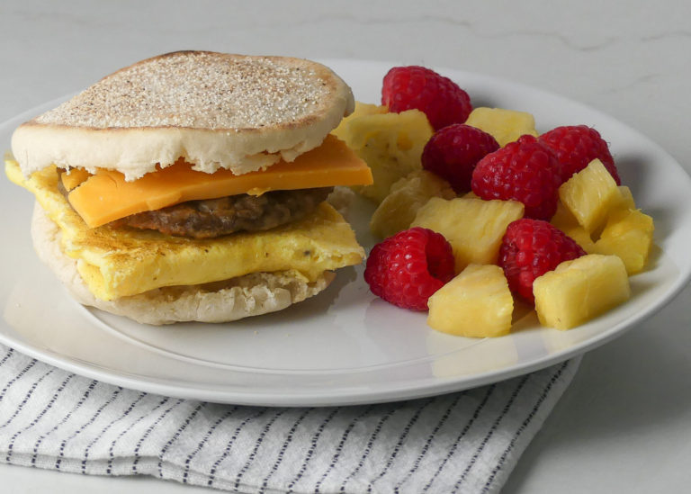 Sausage egg and cheese sandwich on an english muffin with raspberries and pineapple on the side